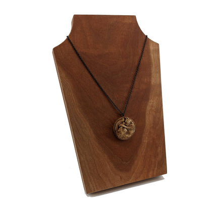 Wooden necklace pendant carved into the shape of a cat curled into a round ball. Hanging from a black stainless steel chain against a cherry wood background.