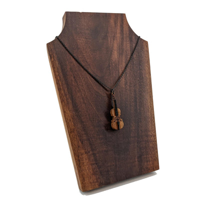 Wooden necklace pendant created in the shape of a miniature cello stand up bass. Made from cherry wood and hanging from a black stainless steel chain against a dark walnut background.