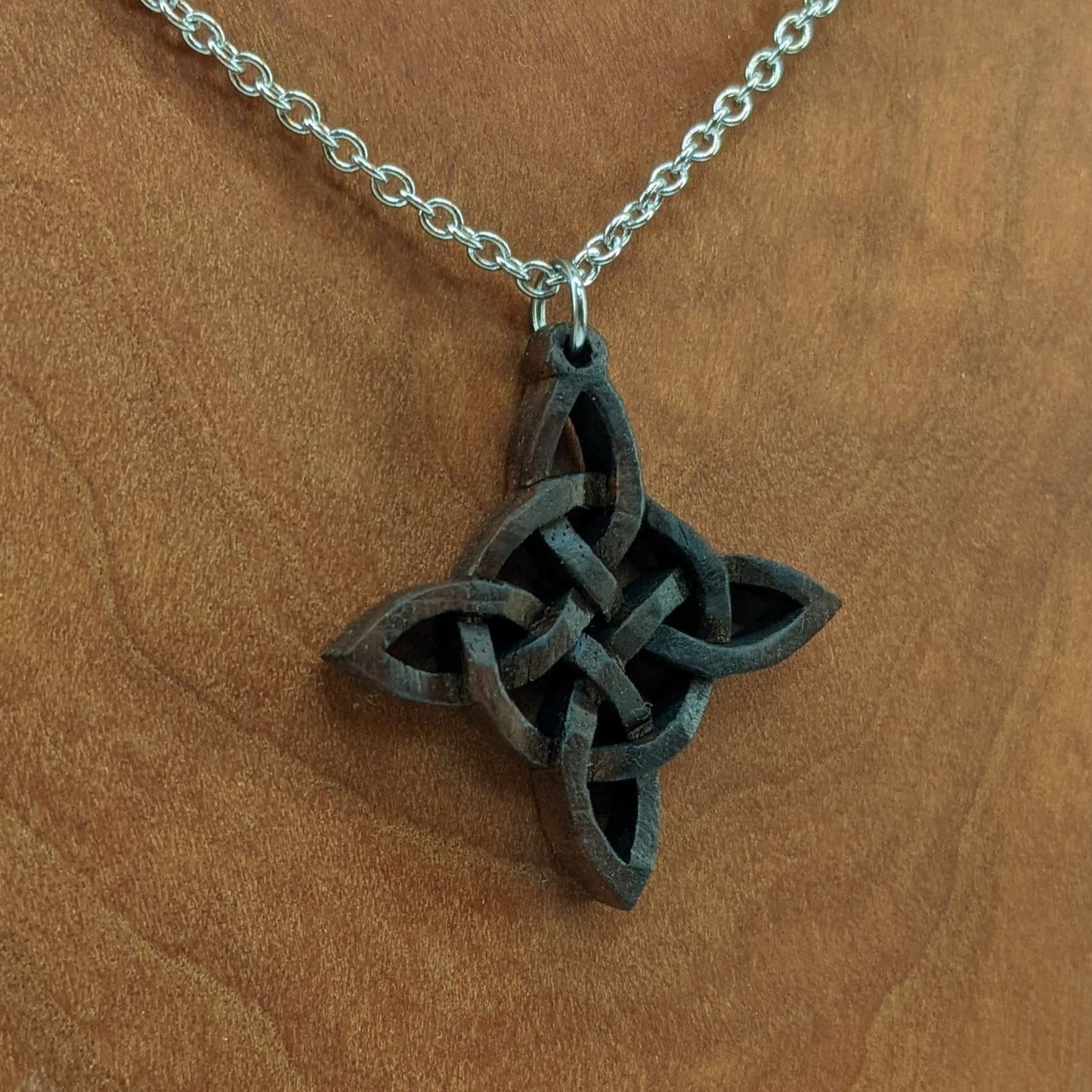 Wooden necklace pendant created in the style of a Celtic woven cross. Made from dark walnut.  Hanging from a silver stainless steel chain against a cherry wood background.