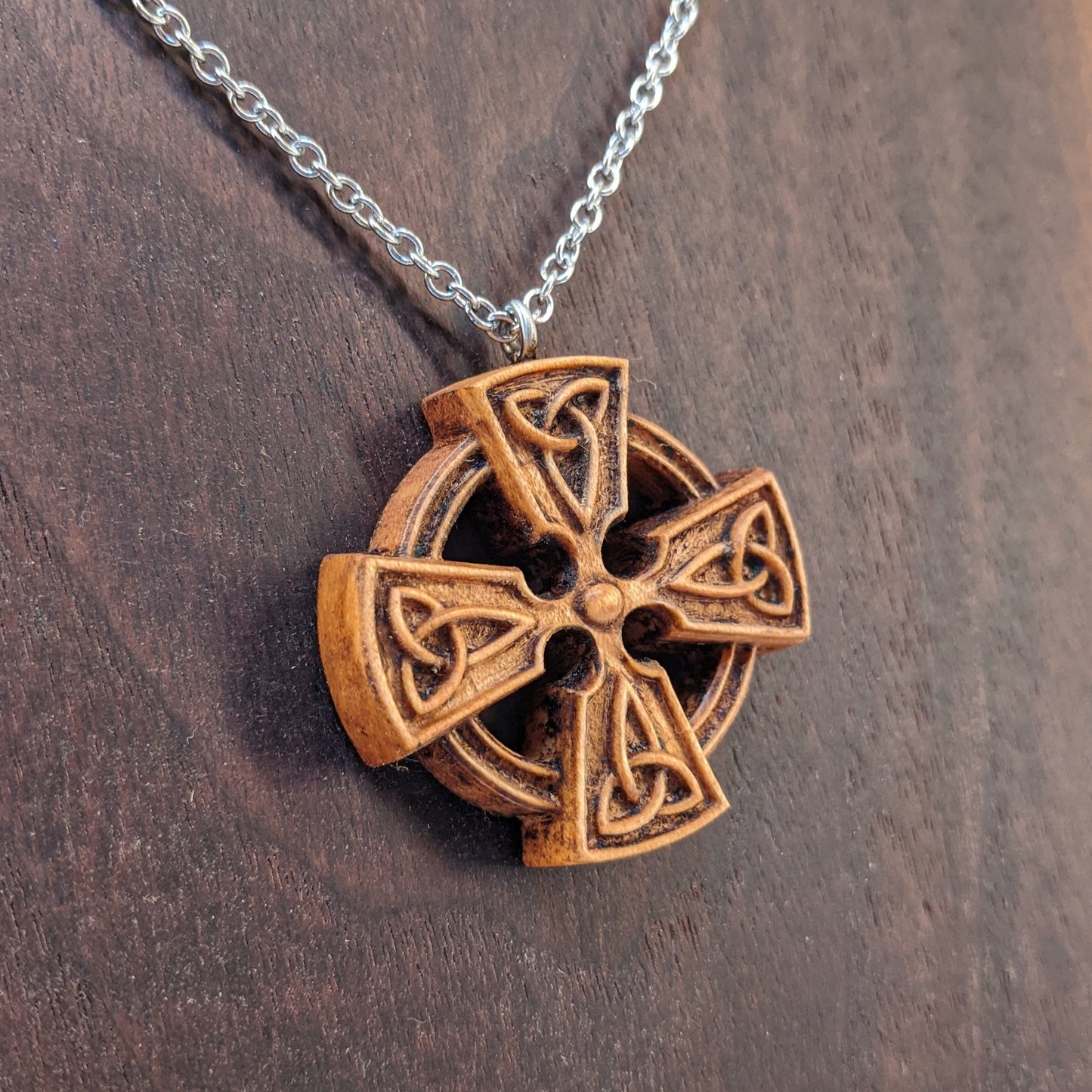 Wooden necklace pendant created in the shape of a Celtic cross. Made from hard maple wood and hanging from a silver stainless steel chain against a dark walnut background.