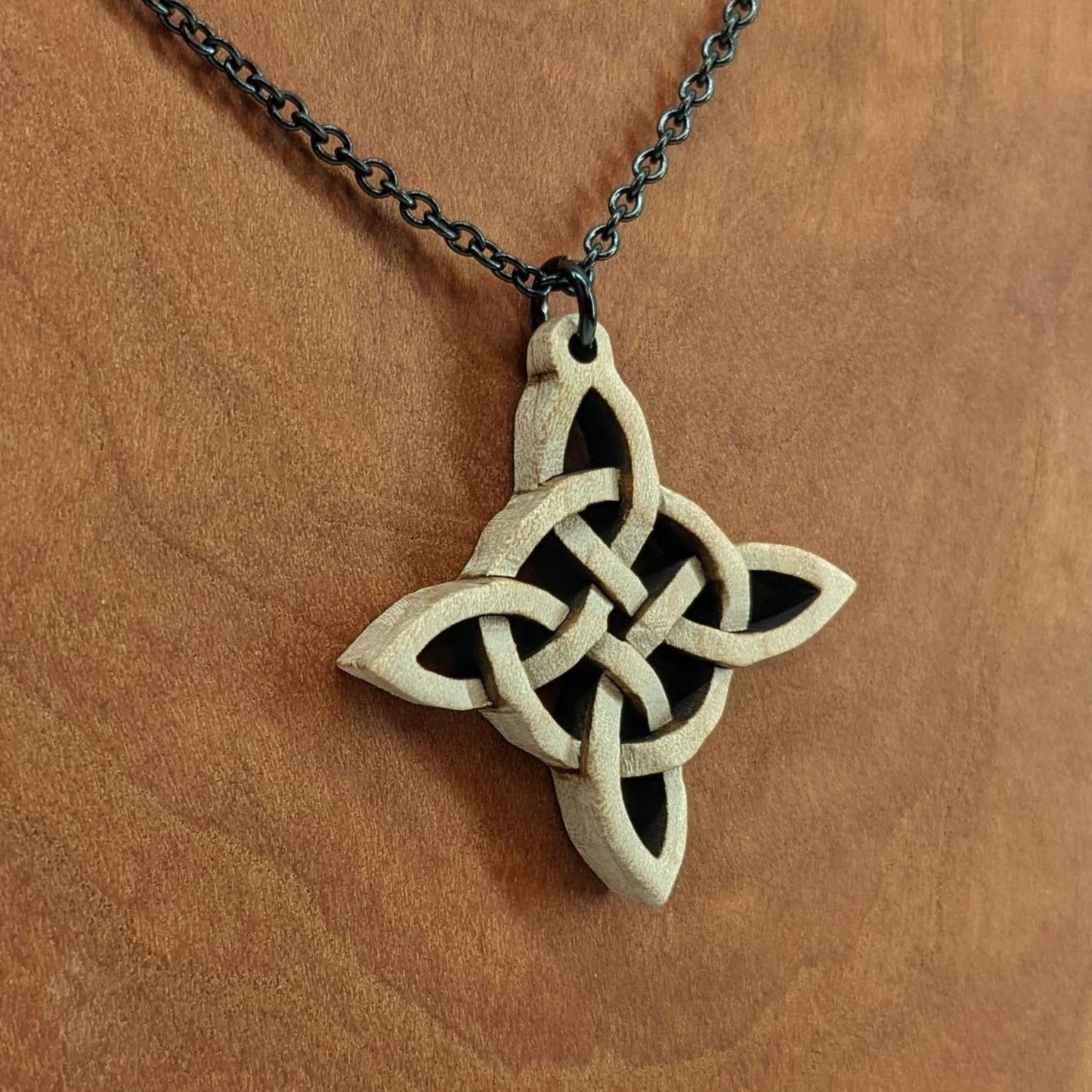 Wooden necklace pendant created in the style of a Celtic woven cross. Made from hard maple wood.  Hanging from a black stainless steel chain against a cherry wood background.