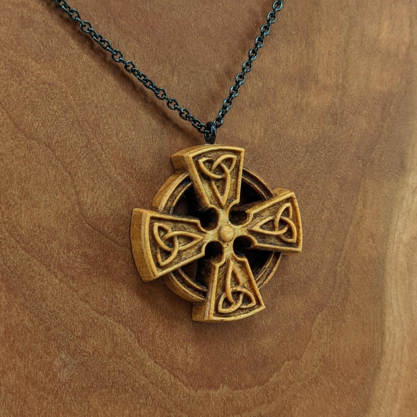 Wooden necklace pendant created in the shape of a Celtic cross. Made from hard maple wood and hanging from a black stainless steel chain against a cherry wood background.