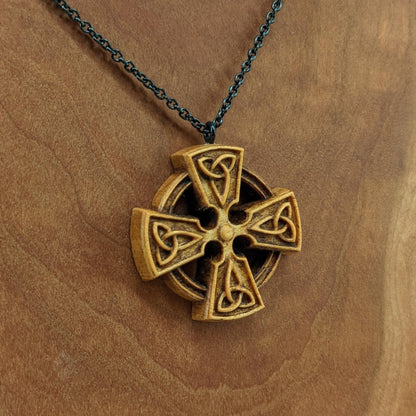 Wooden necklace pendant created in the shape of a Celtic cross. Made from hard maple wood and hanging from a black stainless steel chain against a cherry wood background.