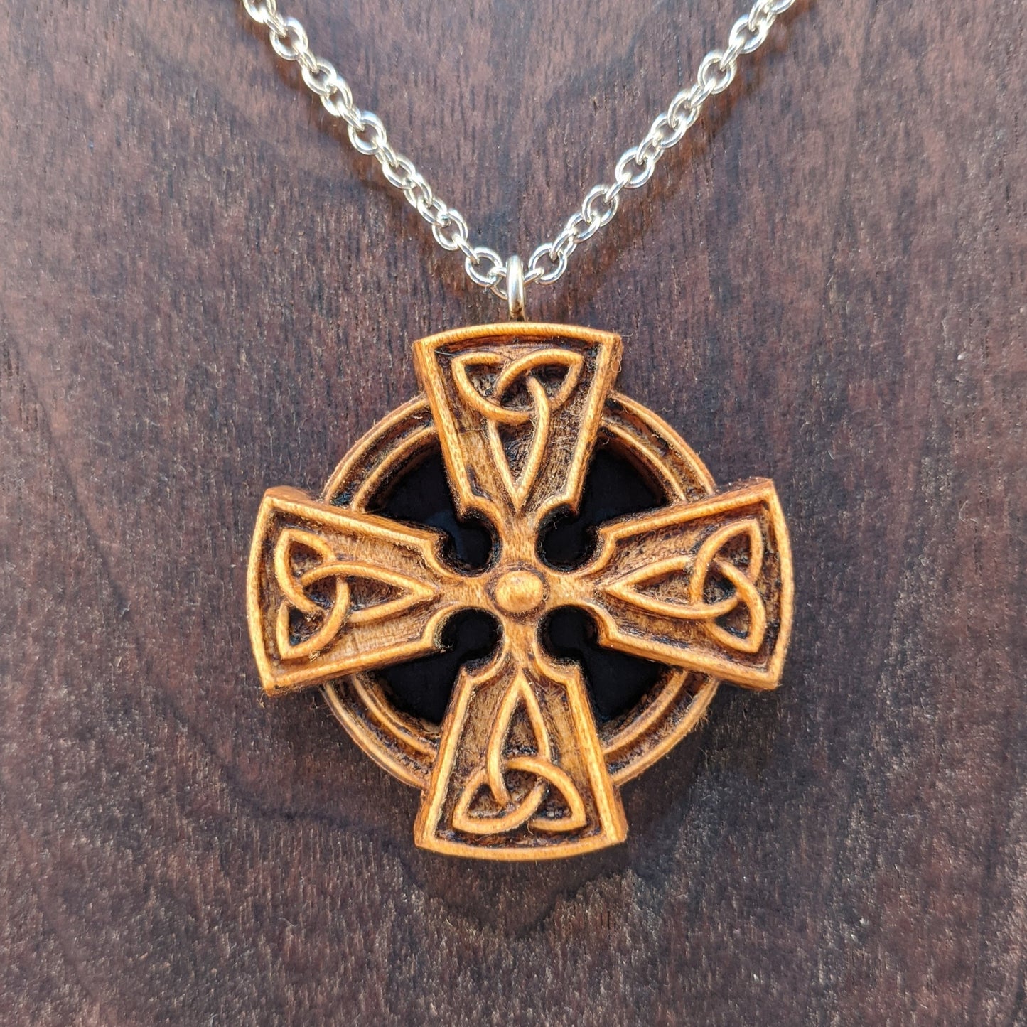 Wooden necklace pendant created in the shape of a Celtic cross. Made from hard maple wood and hanging from a silver stainless steel chain against a dark walnut background.