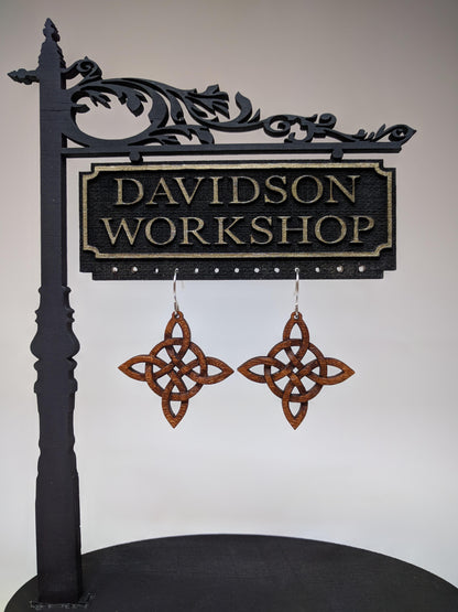 Pair of wooden earrings with silver stainless steel hooks. They are Celtic style crosses, carved in a woven pattern with a circle center. Made from dark walnut hanging from a model Davidson Workshop sign against a white background.