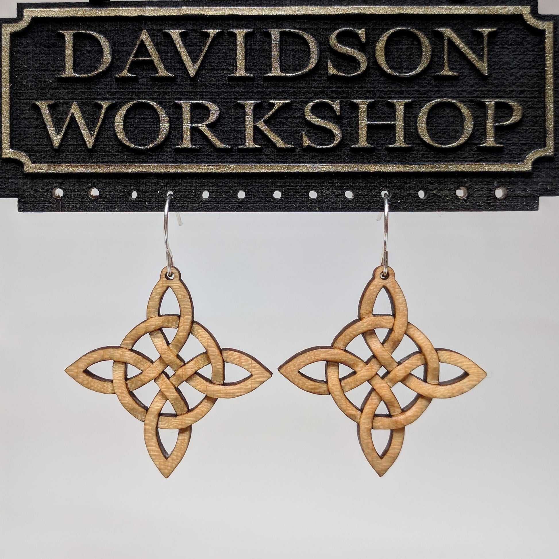 Pair of wooden earrings with silver stainless steel hooks. They are Celtic style crosses, carved in a woven pattern with a circle center. Made from hard maple hanging from a model Davidson Workshop sign against a white background. (Close up View)