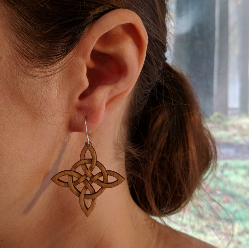 Pair of wooden earrings with silver stainless steel hooks. They are Celtic style crosses, carved in a woven pattern with a circle center. Made from birch wood, being modeled by a women in front of a nature scene.