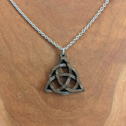 Wooden necklace pendant created in the style of a Celtic woven trinity symbol. Made from dark walnut. Hanging from a silver stainless steel chain against a cherry wood background.
