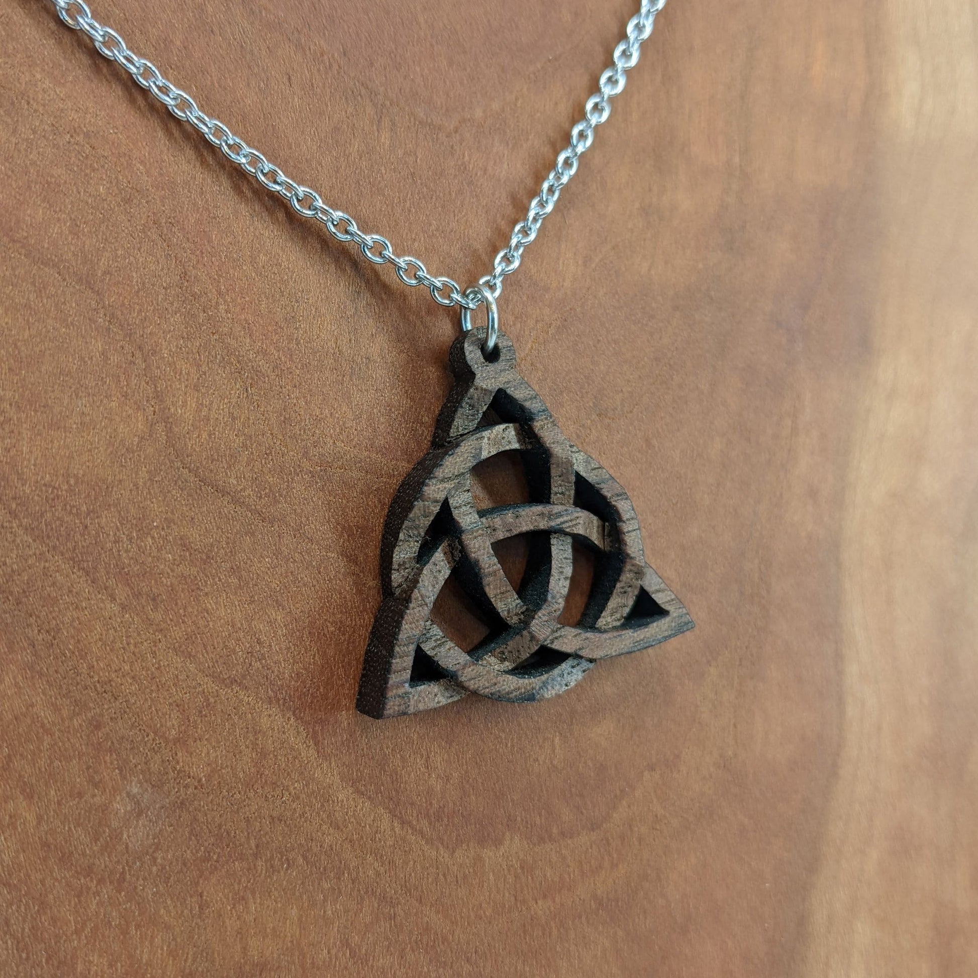 Wooden necklace pendant created in the style of a Celtic woven trinity symbol. Made from dark walnut. Hanging from a silver stainless steel chain against a cherry wood background.