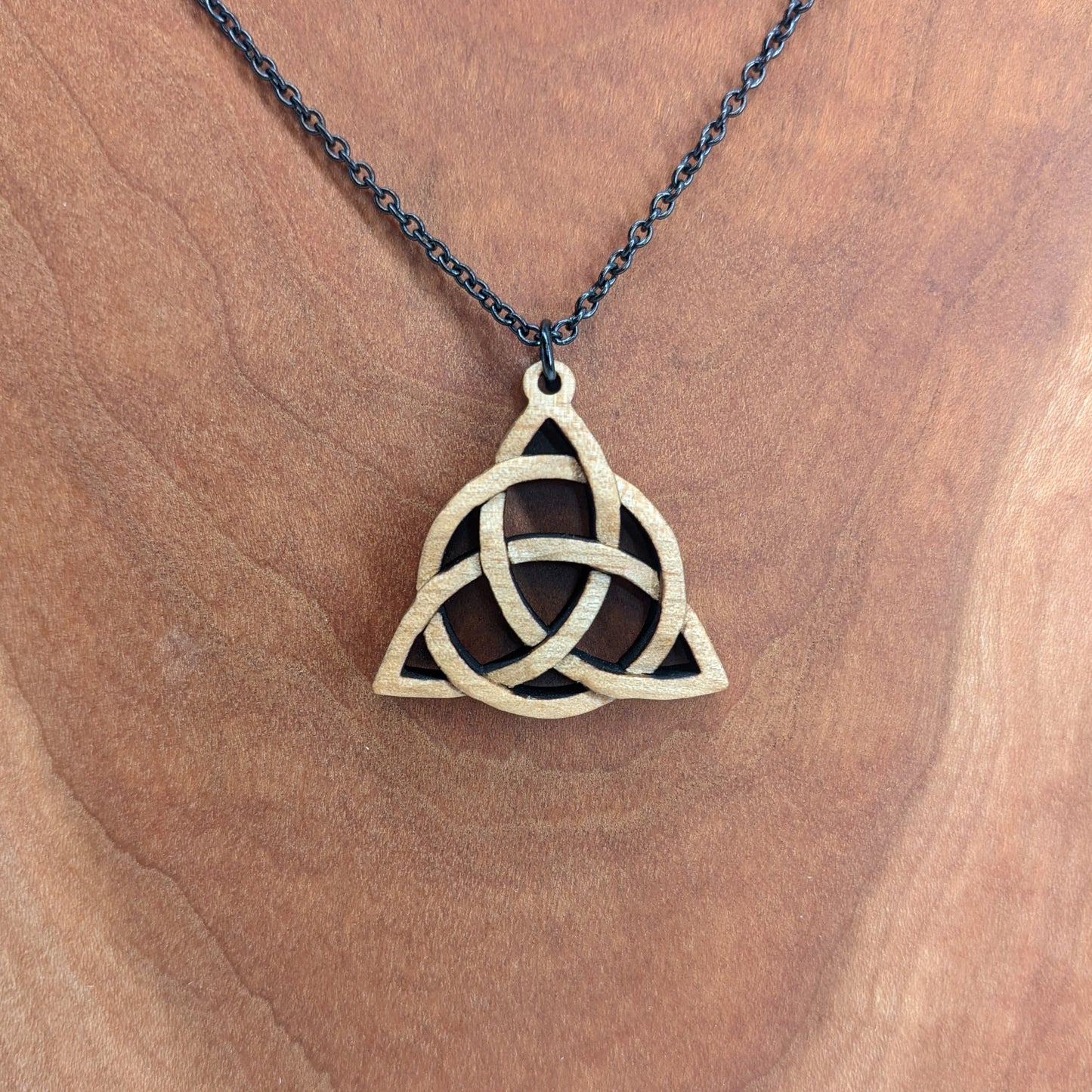 Wooden necklace pendant created in the style of a Celtic woven trinity symbol. Made from hard maple.  Hanging from a black stainless steel chain against a cherry wood background.
