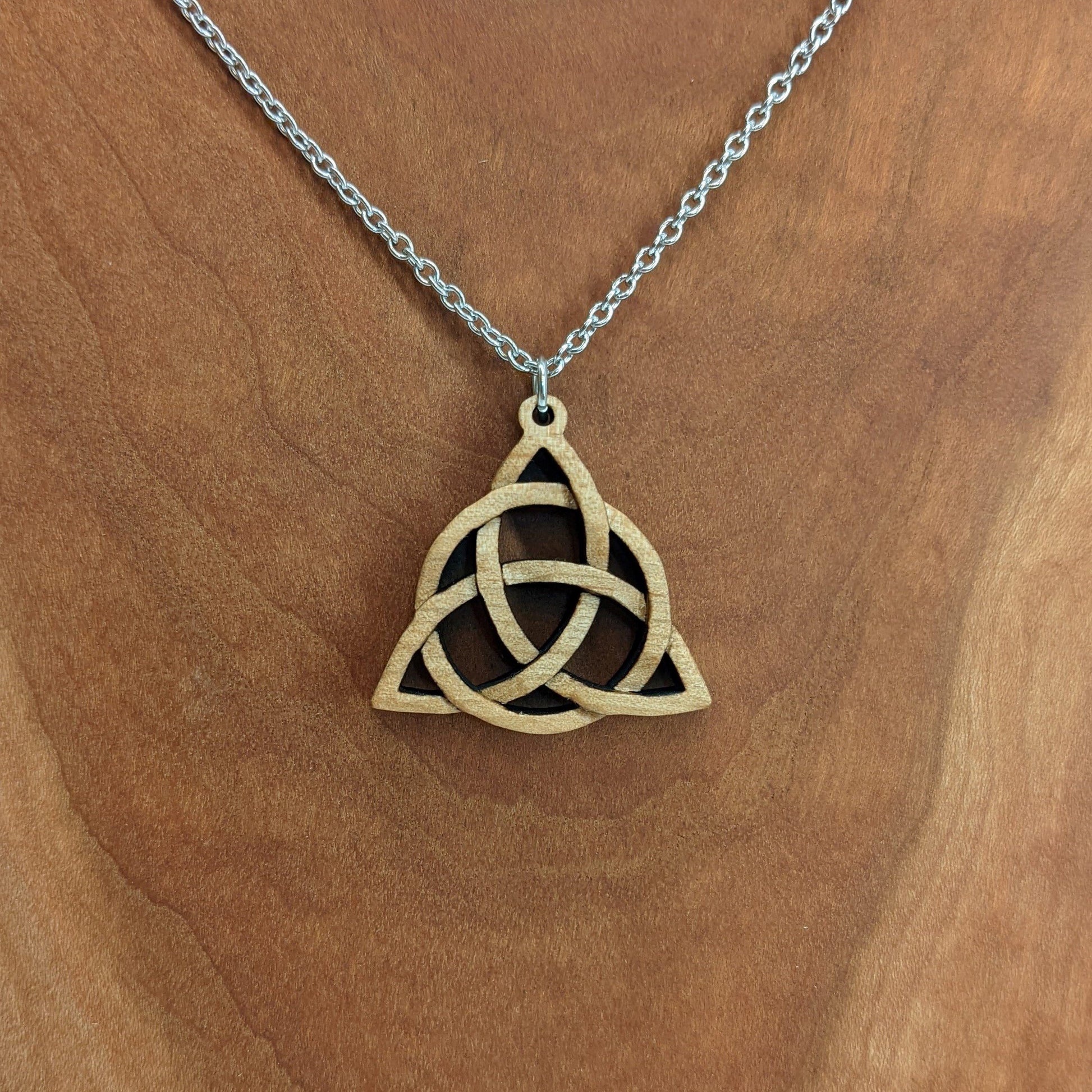 Wooden necklace pendant created in the style of a Celtic woven trinity symbol. Made from hard maple.  Hanging from a silver stainless steel chain against a cherry wood background.