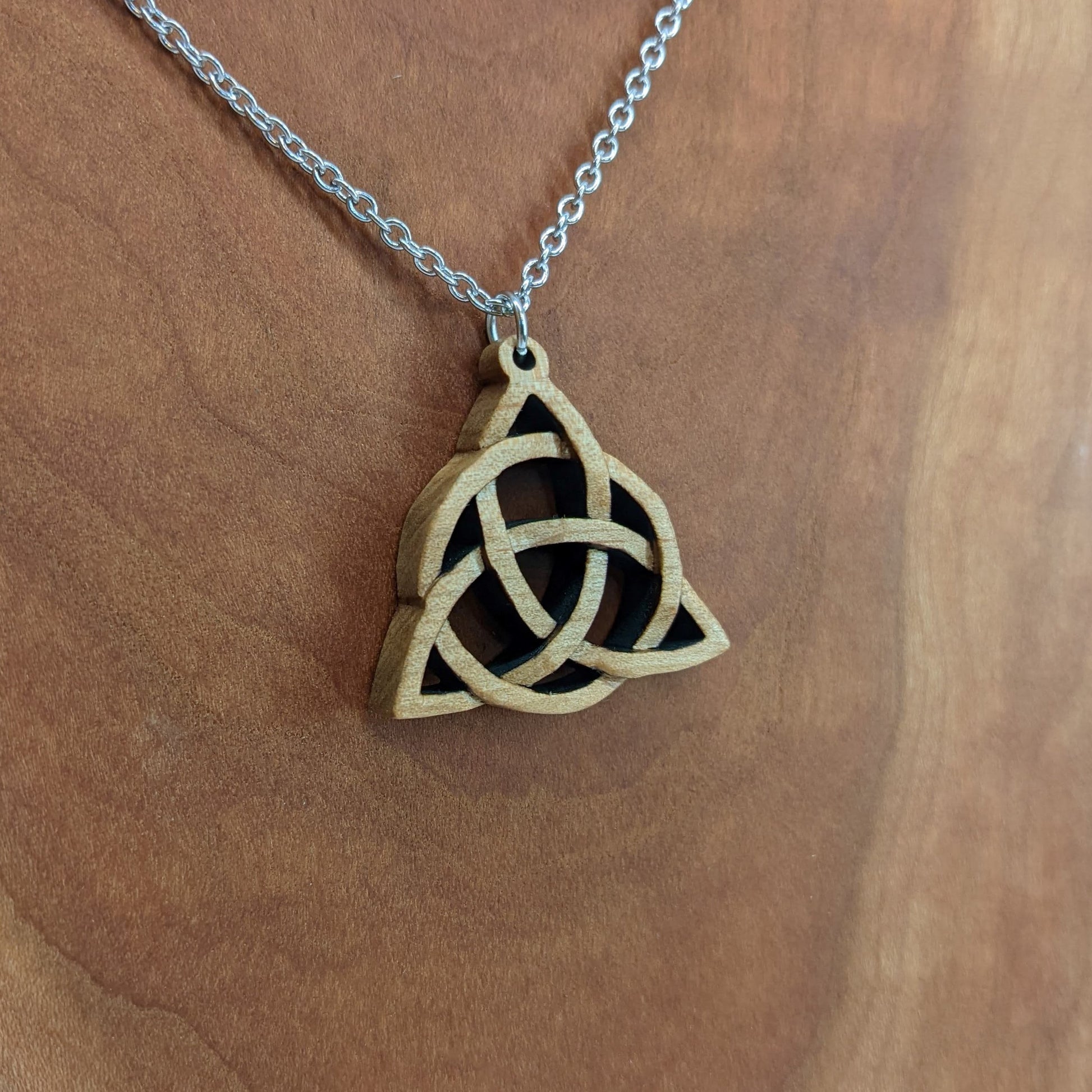 Wooden necklace pendant created in the style of a Celtic woven trinity symbol. Made from hard maple.  Hanging from a silver stainless steel chain against a cherry wood background.