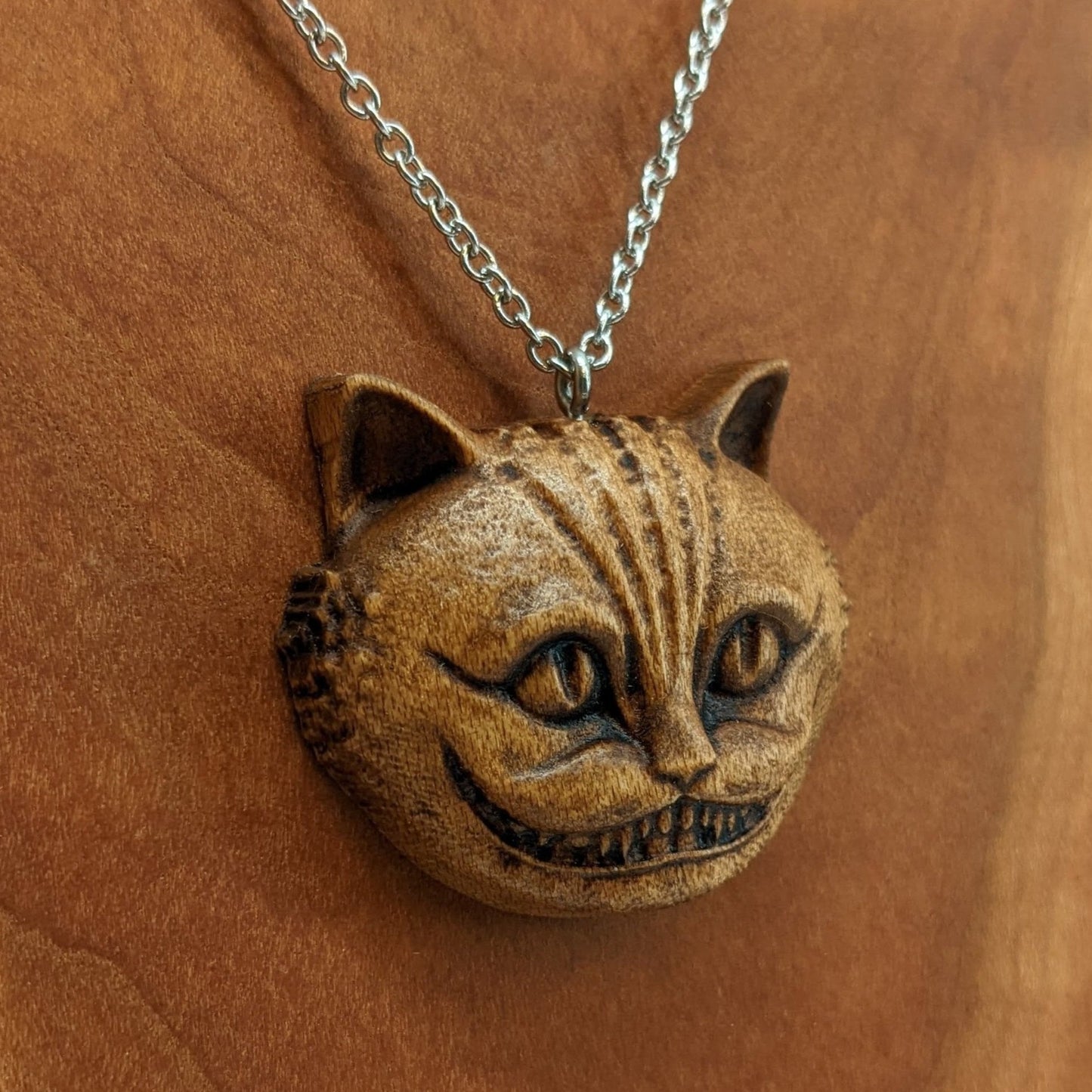 Wooden necklace pendant carved in the shape of a Cheshire cat head. Hanging from a silver stainless steel chain against a cherry wood background.