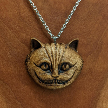 Wooden necklace pendant carved in the shape of a Cheshire cat head. Hanging from a silver stainless steel chain against a cherry wood background.