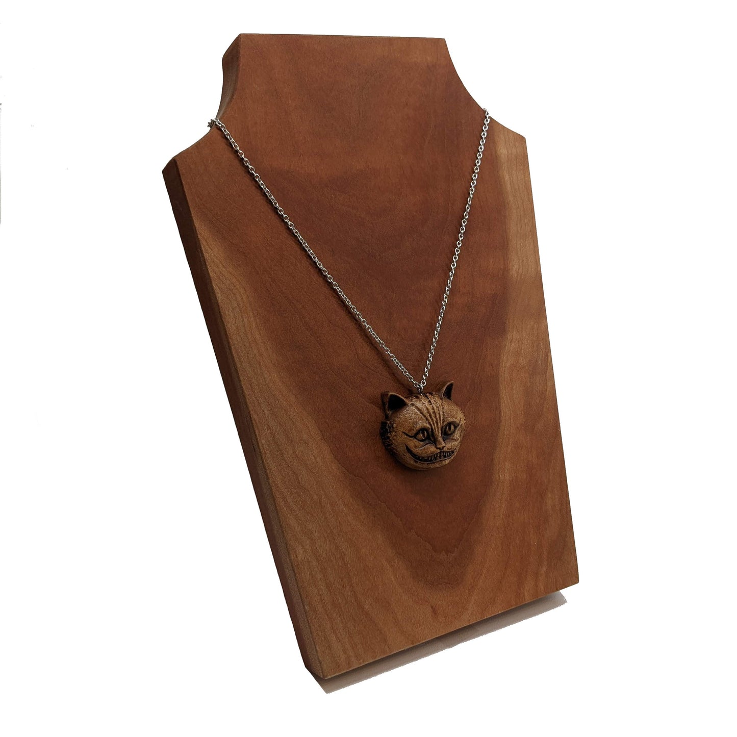 Wooden necklace pendant carved in the shape of a Cheshire cat head. Hanging from a silver stainless steel chain against a cherry wood display.