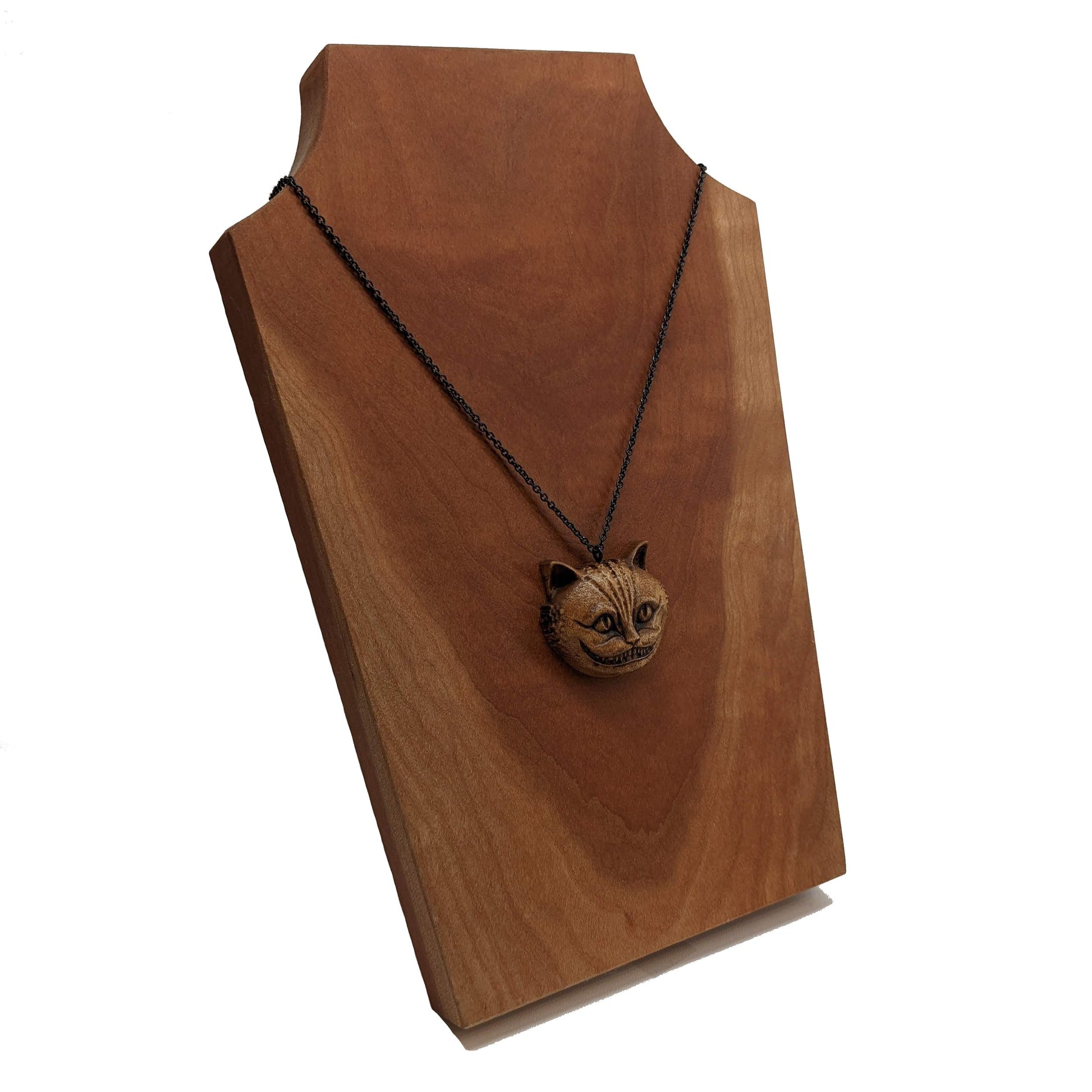 Wooden necklace pendant carved in the shape of a Cheshire cat head. Hanging from a black stainless steel chain against a cherry wood display.