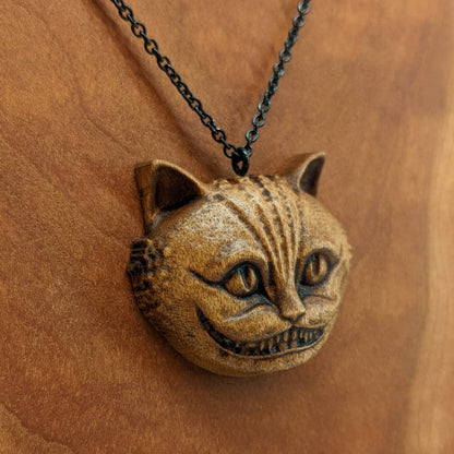 Wooden necklace pendant carved in the shape of a Cheshire cat head. Hanging from a black stainless steel chain against a cherry wood background.