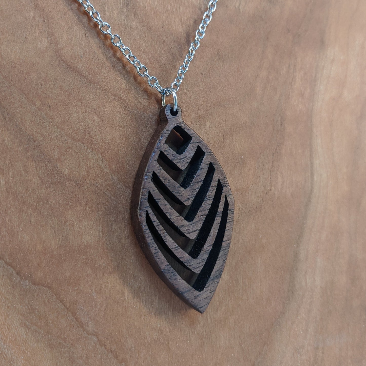 Wooden necklace pendant cut into a teardrop shape with a chevron pattern cut throughout it. Made from walnut and hanging from a silver stainless steel chain against a cherry background.