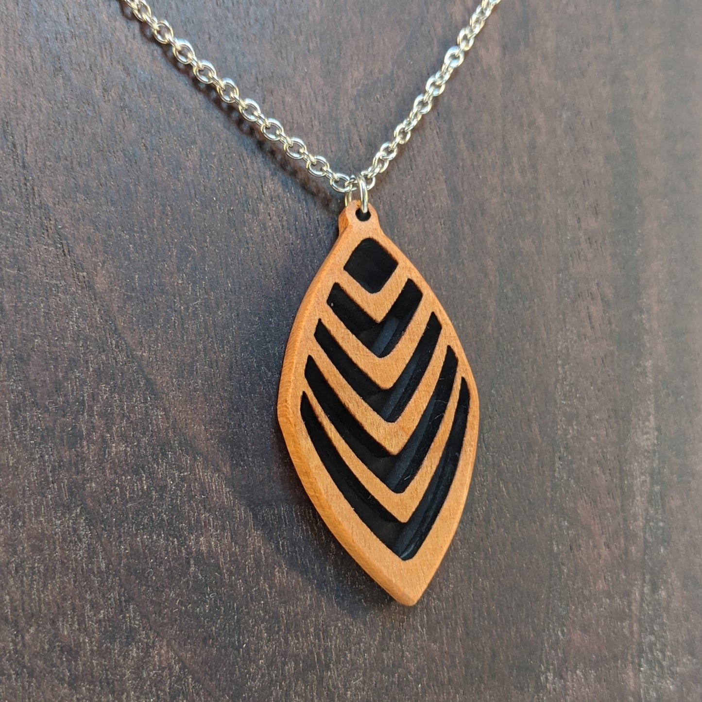 Wooden necklace pendant cut into a teardrop shape with a chevron pattern cut throughout it. Made from cherry and hanging from a silver stainless steel chain against a dark walnut background.