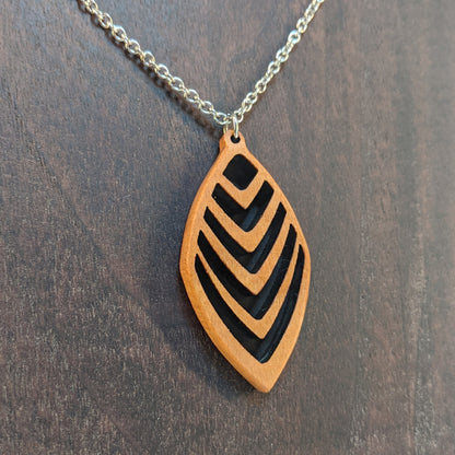 Wooden necklace pendant cut into a teardrop shape with a chevron pattern cut throughout it. Made from cherry and hanging from a silver stainless steel chain against a dark walnut background.