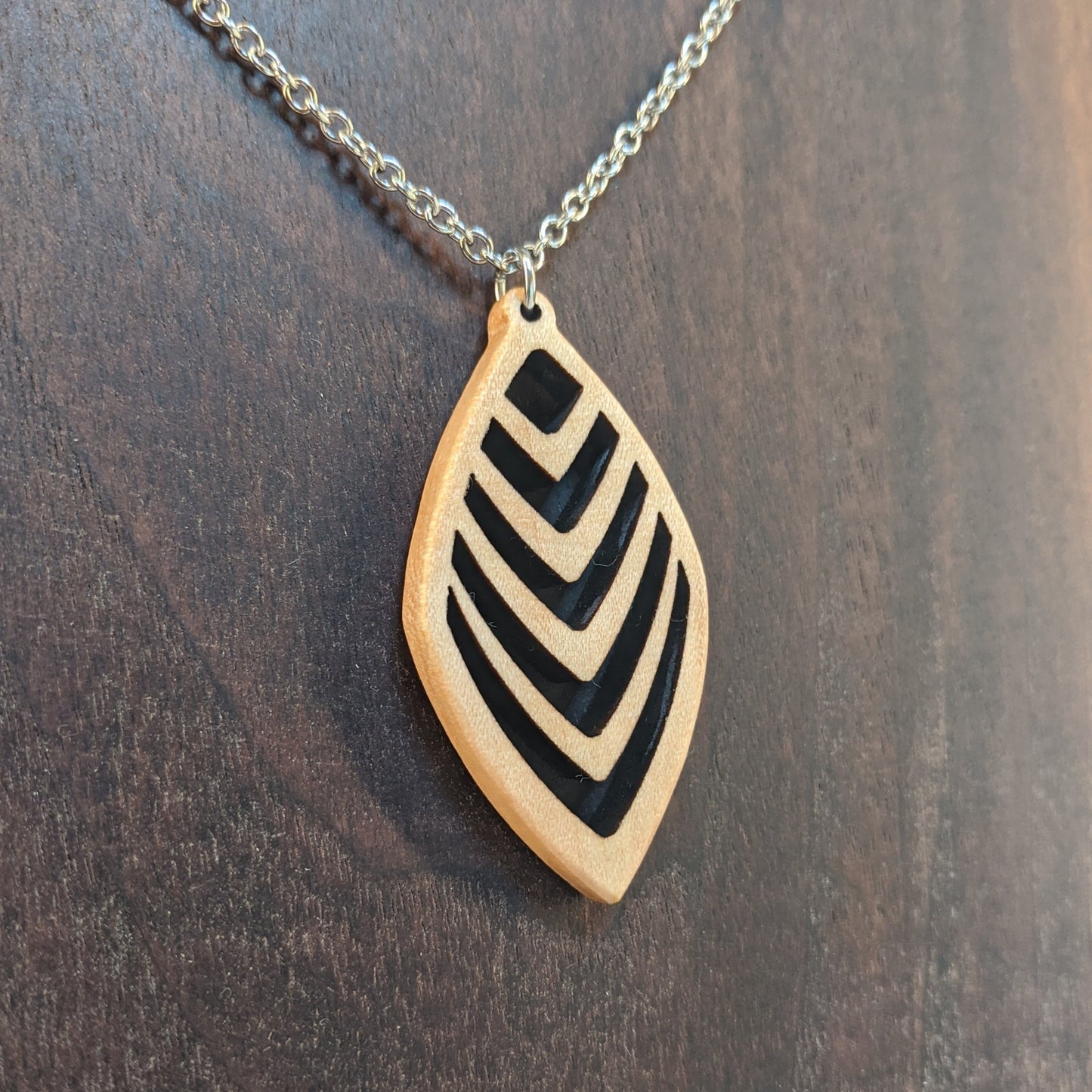 Wooden necklace pendant cut into a teardrop shape with a chevron pattern cut throughout it. Made from hard maple and hanging from a silver stainless steel chain against a dark walnut background.