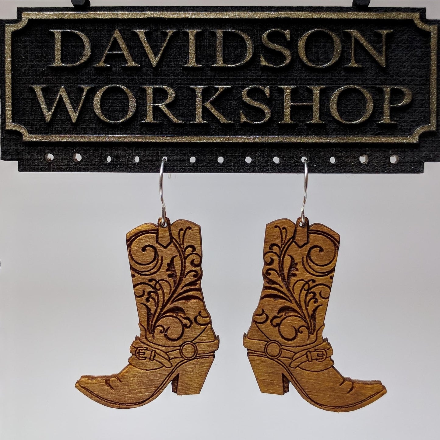 Pair of wooden earrings with silver stainless steel hooks. They are brown cowboy boots with filigree patterns engraved up the sides. Made from birch wood hanging from a model Davidson Workshop sign against a white background. (Close up View)