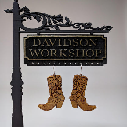 Pair of wooden earrings with silver stainless steel hooks. They are brown cowboy boots with filigree patterns engraved up the sides. Made from birch wood hanging from a model Davidson Workshop sign against a white background.