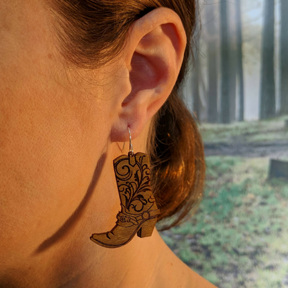 Pair of wooden earrings with silver stainless steel hooks. They are brown cowboy boots with filigree patterns engraved up the sides. Made from birch wood, being modeled by a women in front of a nature scene.