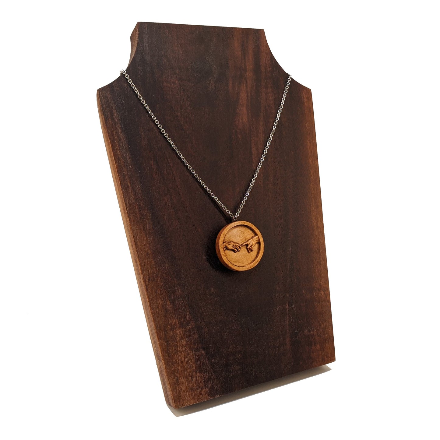 Round wooden necklace pendant carved in the shape of 2 hands reaching out to touch each other. The hands are carved from light maple wood. Hanging from a silver stainless steel chain against a dark walnut background.