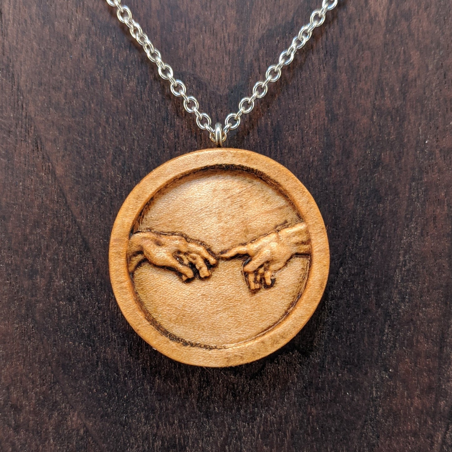 Round wooden necklace pendant carved in the shape of 2 hands reaching out to touch each other. The hands are carved from light maple wood. Hanging from a silver stainless steel chain against a dark walnut background.