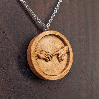 Round wooden necklace pendant carved in the shape of 2 hands reaching out to touch each other. The hands are carved from light maple wood. Hanging from a silver stainless steel chain against a dark walnut background.