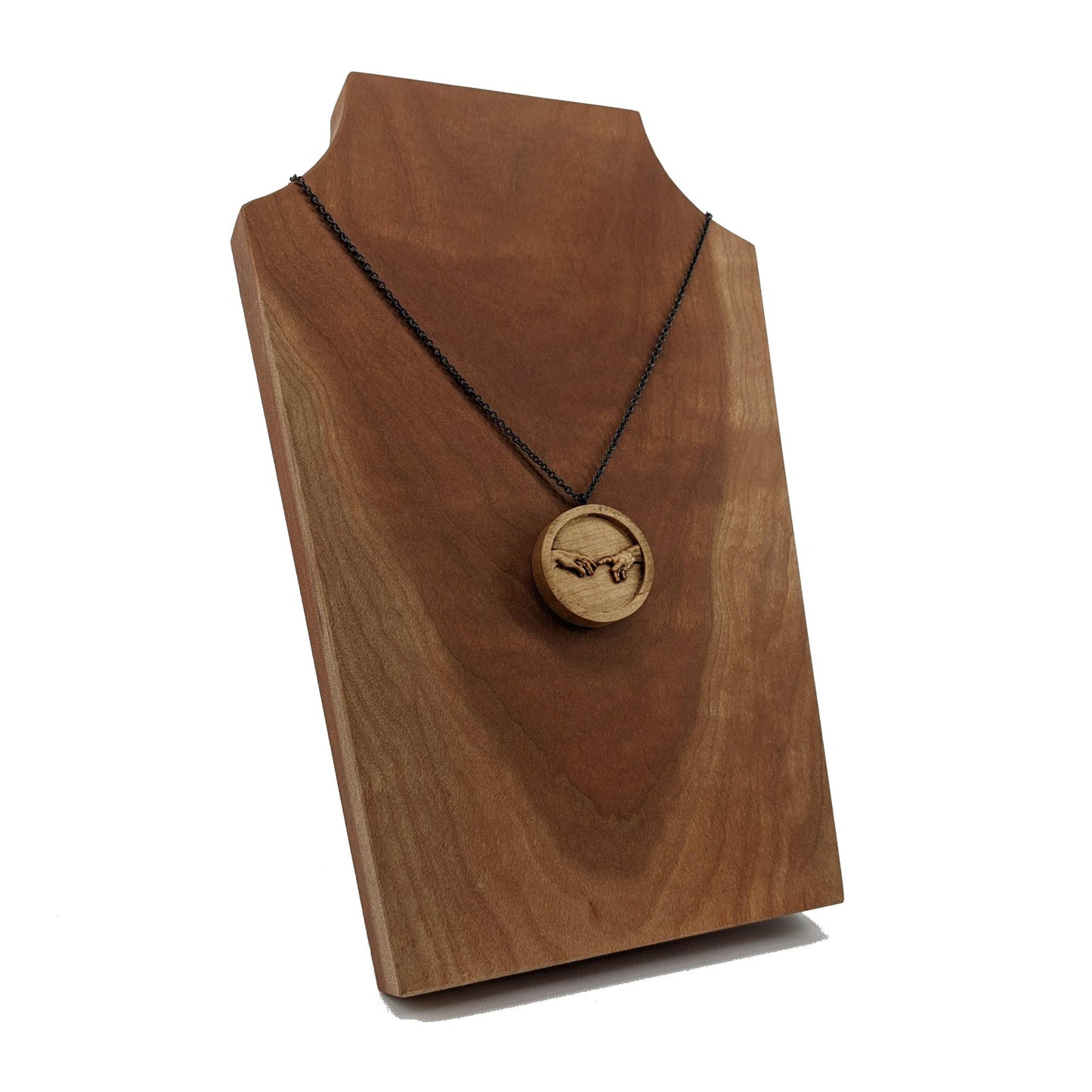 Round wooden necklace pendant carved in the shape of 2 hands reaching out to touch each other. The hands are carved from light maple wood. Hanging from a silver stainless steel chain against a dark walnut display.