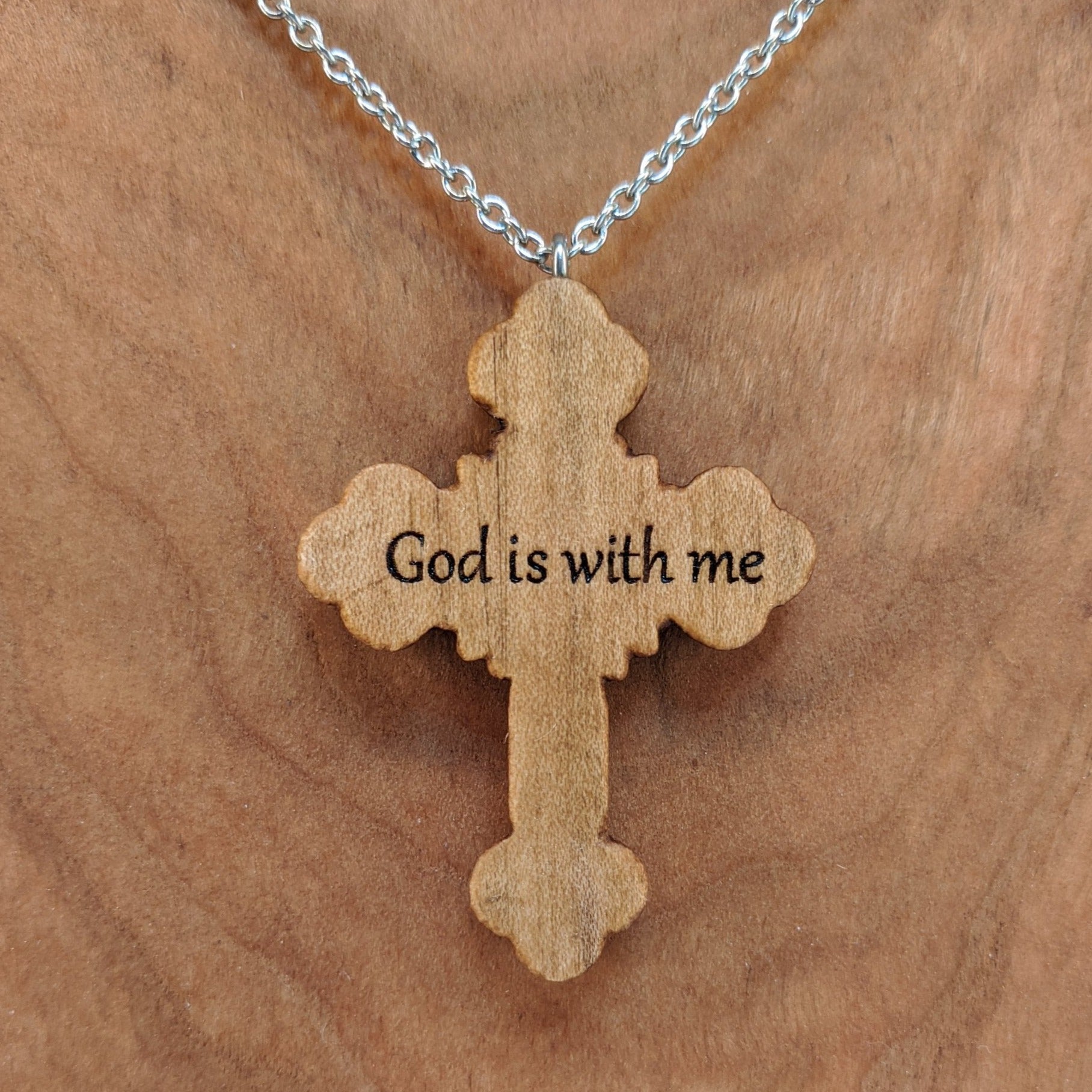 Wooden necklace pendant carved in the shape of an ornate cross. The back is engraved with personalized wording. Made from hard maple wood and hanging from a silver stainless steel chain against a cherry wood background.