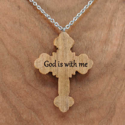 Wooden necklace pendant carved in the shape of an ornate cross. The back is engraved with personalized wording. Made from hard maple wood and hanging from a silver stainless steel chain against a cherry wood background.