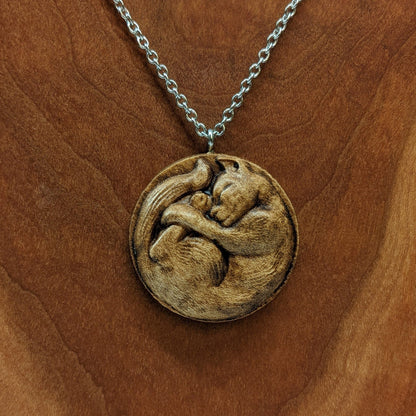 Wooden necklace pendant carved into the shape of a cat curled into a round ball. Hanging from a silver stainless steel chain against a cherry wood background.