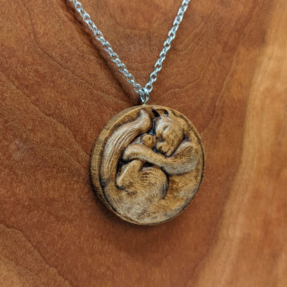 Wooden necklace pendant carved into the shape of a cat curled into a round ball. Hanging from a silver stainless steel chain against a cherry wood background.