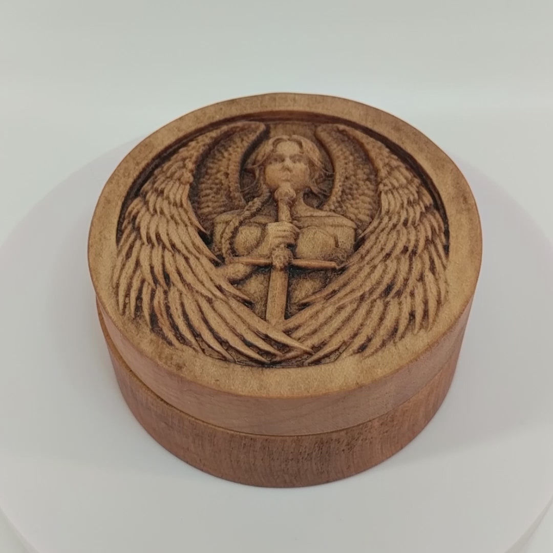 Round wooden box 3D carved with a warrior angel with a serious face and a sword straight down in front. She has parted bangs and large intricately layered wings with sharp tips. Made from hard maple wood stained brown against a white background rotating on a table.