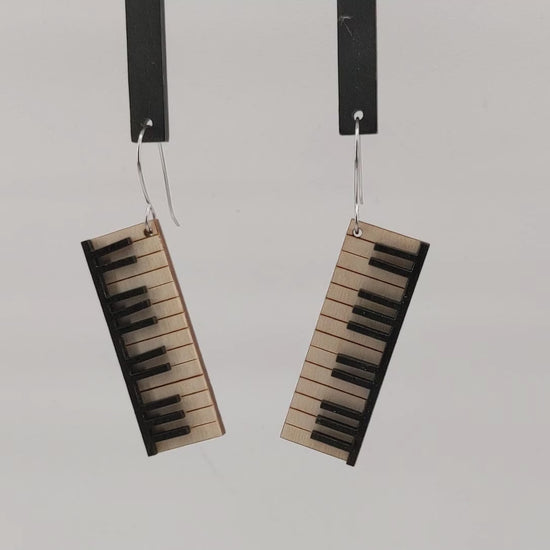 Pair of wooden earrings with silver stainless steel hooks. They are pale natural finished miniature keyboards with painted black keys. Made from birch wood hanging and rotating against a white background.