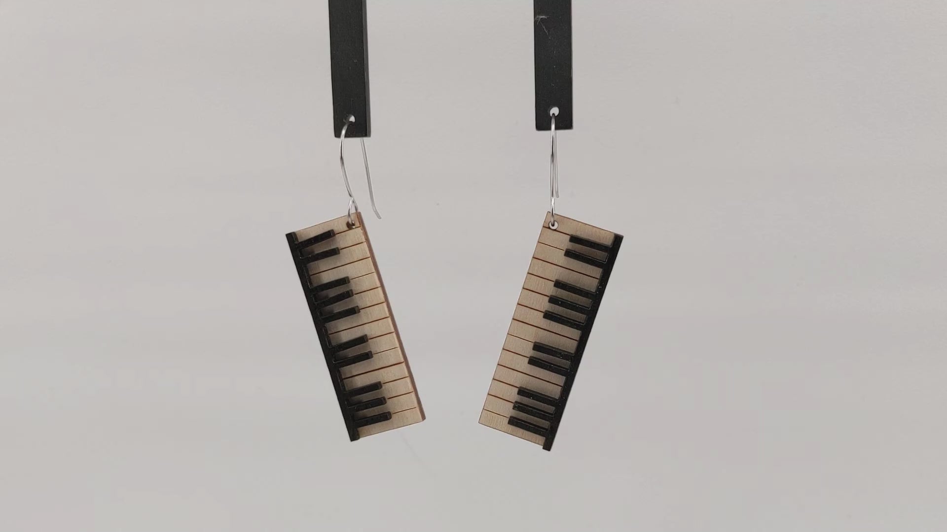 Pair of wooden earrings with silver stainless steel hooks. They are pale natural finished miniature keyboards with painted black keys. Made from birch wood hanging and rotating against a white background.