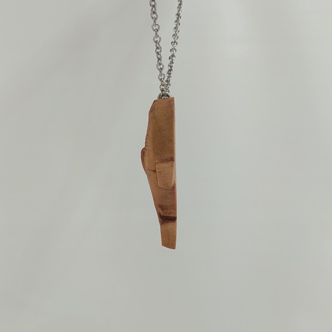 Wooden pendant necklace carved in the shape of great white shark. Made from hard maple wood. The wood shark hangs from its mouth on a silver stainless steel chain against a white background rotating on a table.