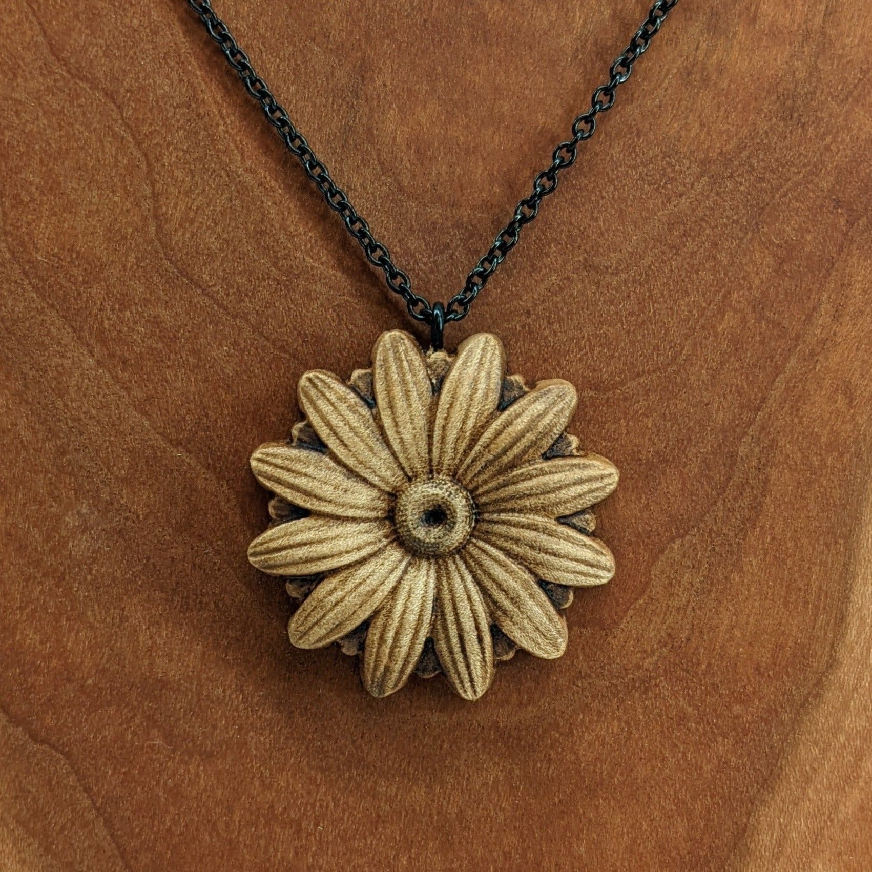 Wooden necklace pendant carved into the shape of a daisy flower. Hanging from a black stainless steel chain against a cherry wood background.