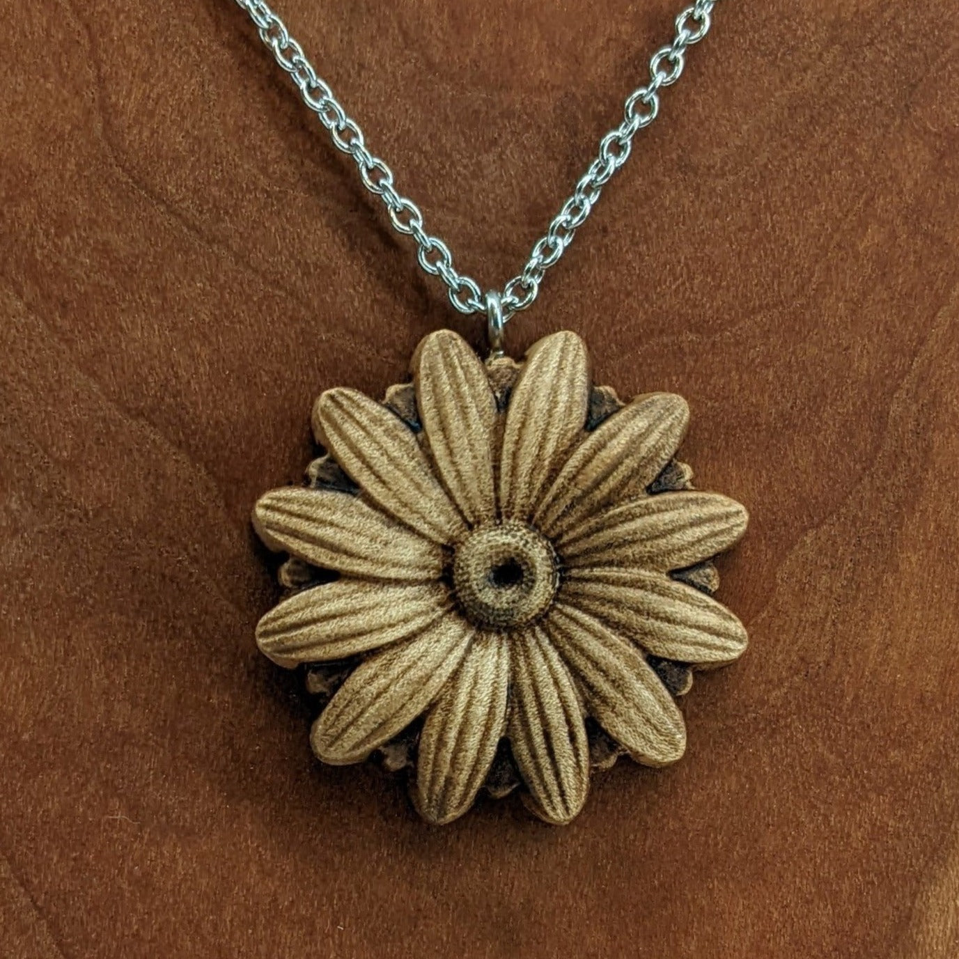 Wooden necklace pendant carved into the shape of a daisy flower. Hanging from a silver stainless steel chain against a cherry wood background.