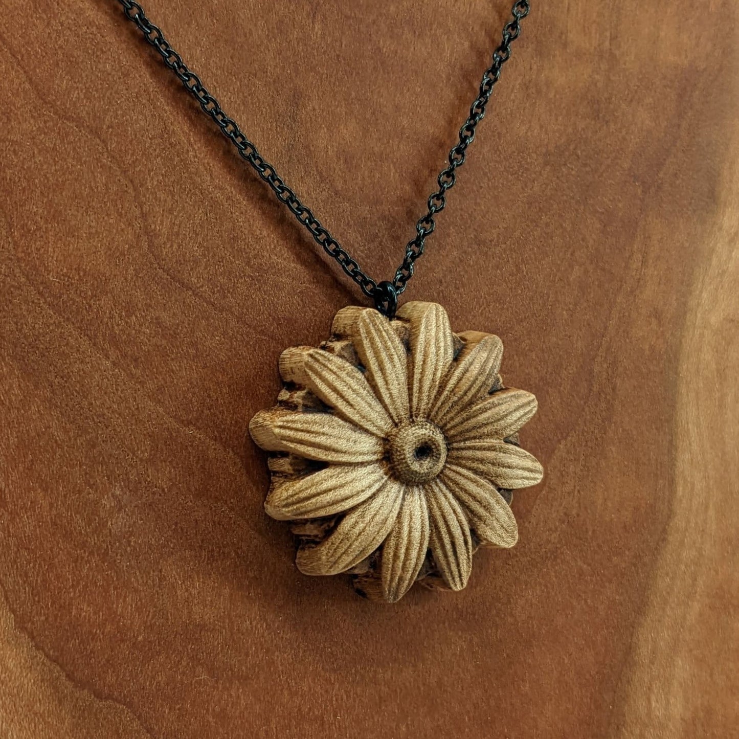 Wooden necklace pendant carved into the shape of a daisy flower. Hanging from a black stainless steel chain against a cherry wood background.