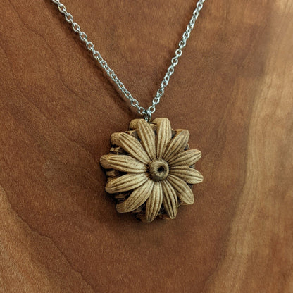 Wooden necklace pendant carved into the shape of a daisy flower. Hanging from a silver stainless steel chain against a cherry wood background.