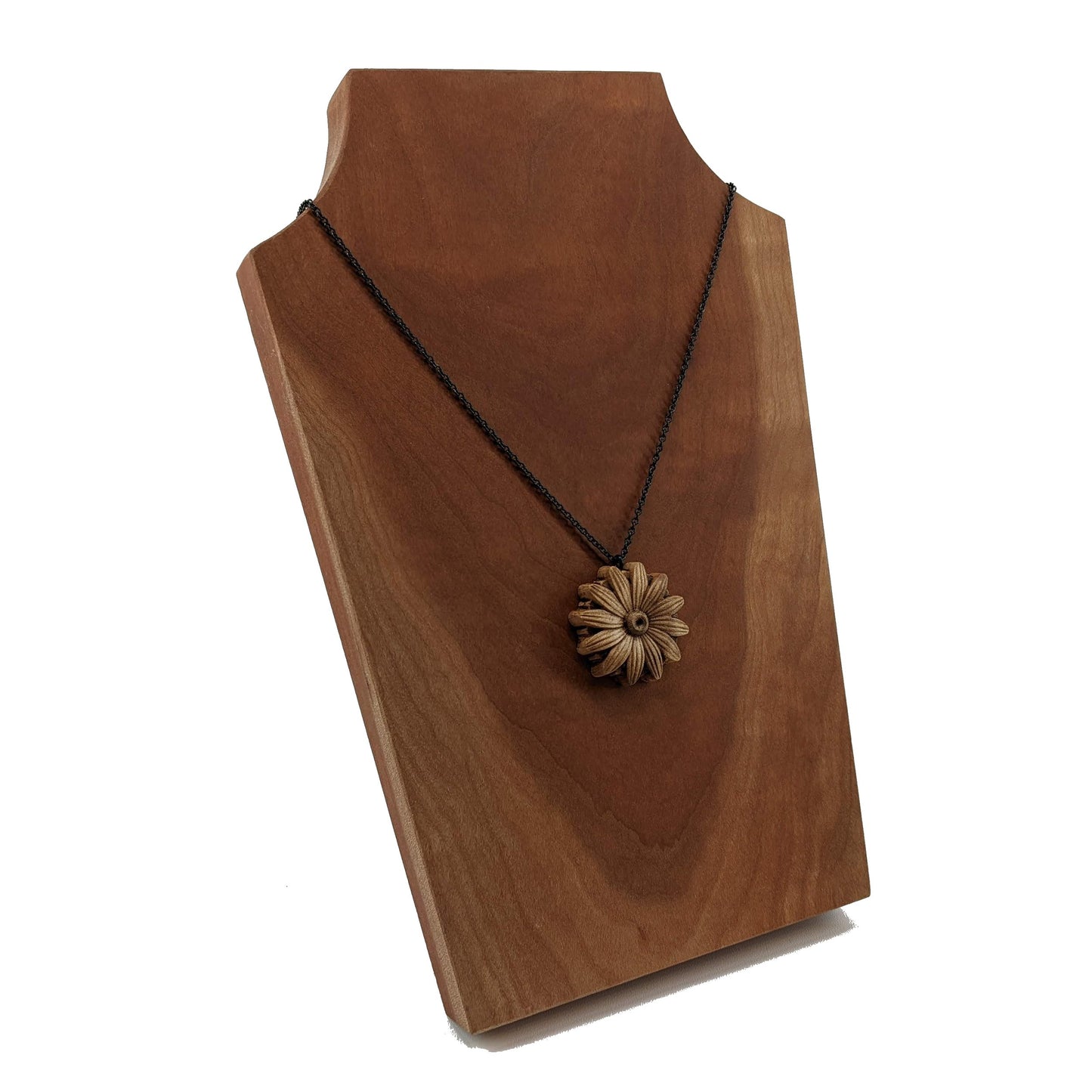 Wooden necklace pendant carved into the shape of a daisy flower. Hanging from a black stainless steel chain against a cherry wood background.