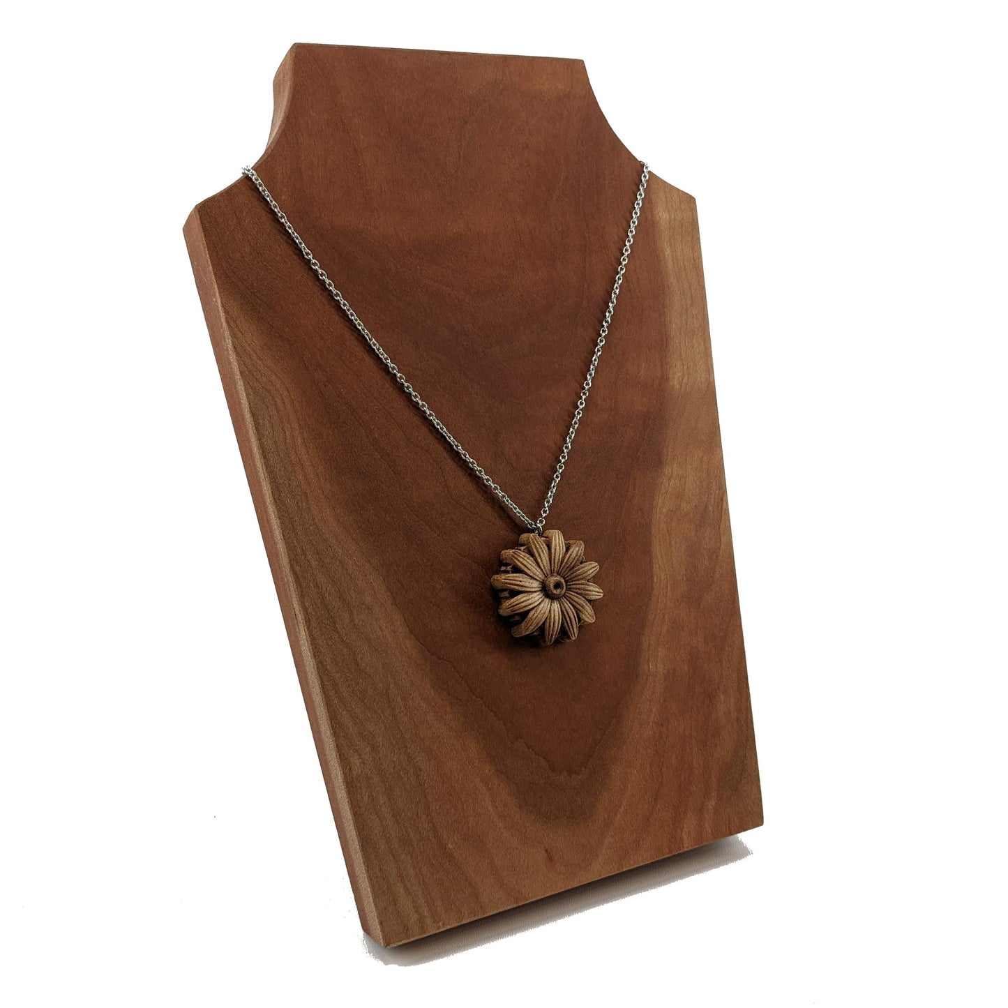 Wooden necklace pendant carved into the shape of a daisy flower. Hanging from a silver stainless steel chain against a cherry wood background.