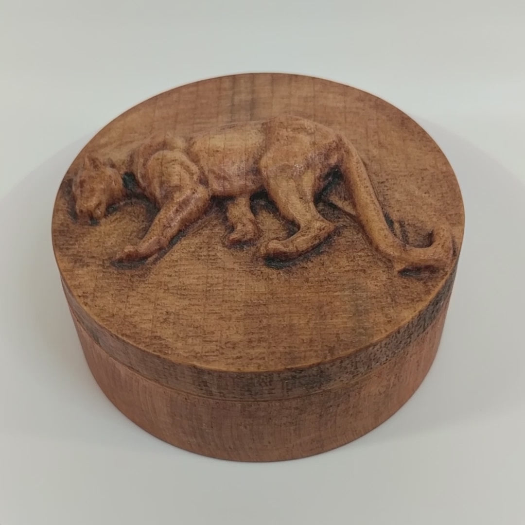 Round wooden box 3D carved with a mountain lion standing broadside to the left. It has a sleek body with its head bent toward the ground and a long tail that hangs to its feet. Made from hard maple wood stained brown against a white background rotating on a table.