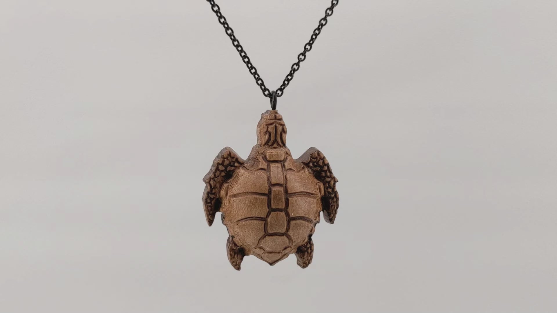 Wooden necklace pendant carved in the shape of a sea turtle. Made from hard maple wood. Hanging and rotating from a black stainless steel chain against a white background.