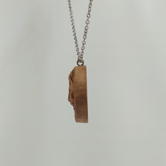 Wooden necklace pendant carved in the shape of a stern lion face surrounded by a rope border. Hanging from a silver stainless steel chain against a white background rotating on a table. 