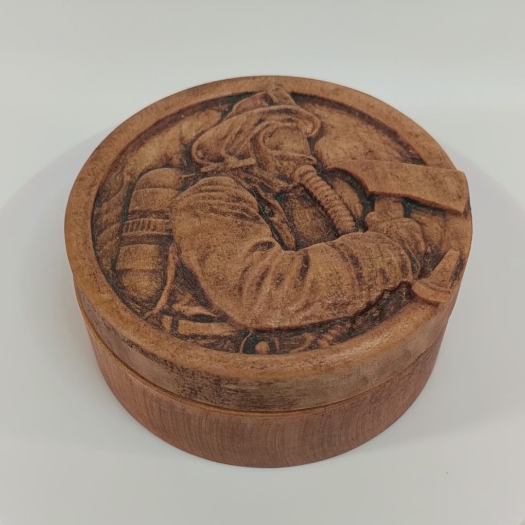 Round wooden box 3D carved with a firefighter in full gear with hat, oxygen tank, mask on and axe in hand within a fire hose border in front of a background of flames. Made from hard maple wood with a brown stain against a white background rotating on a table. 